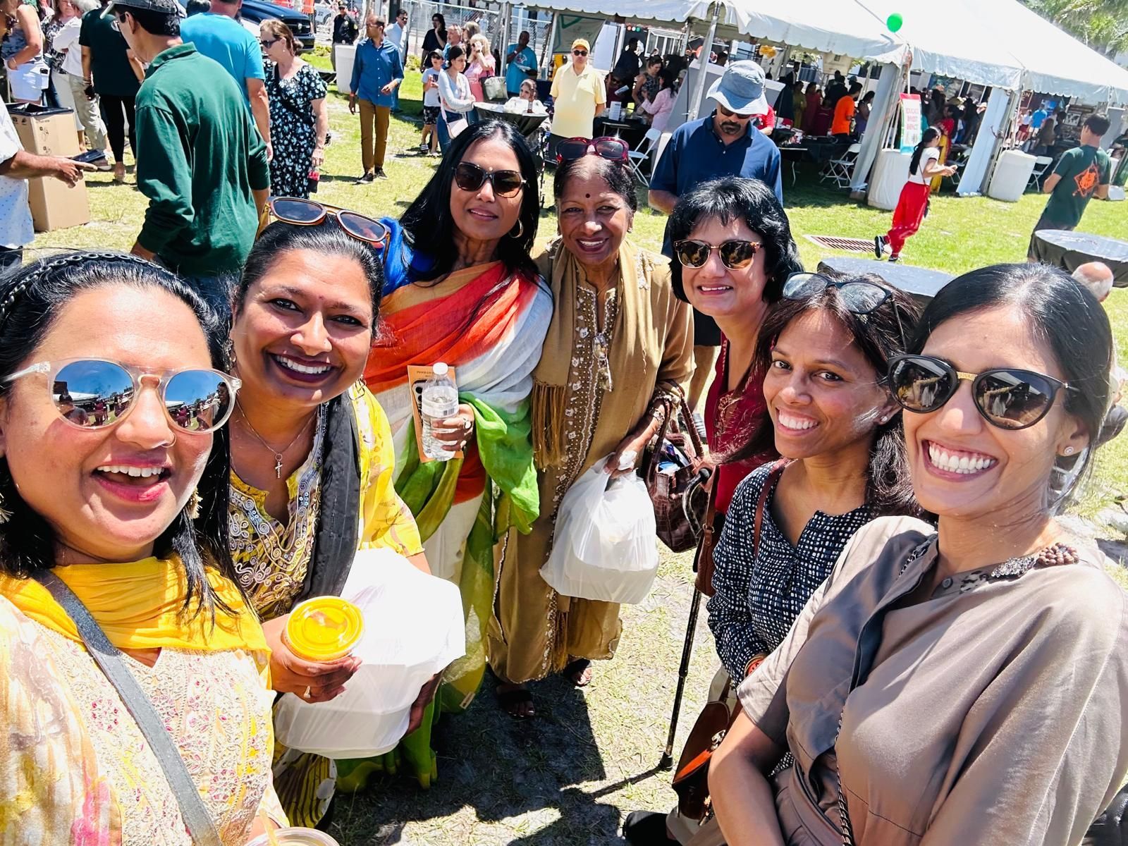 Group of women smiling, taking a selfie at an outdoor festival. Sunny day, green grass, food stalls in the background.