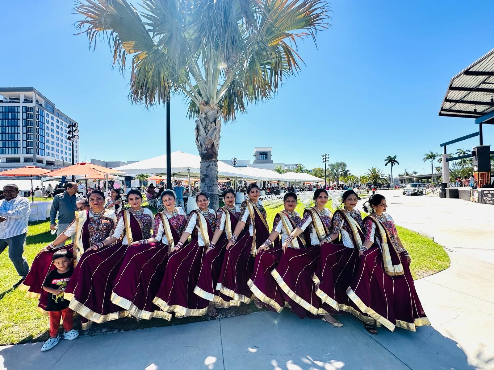 Group of dancers in maroon saris with gold trim, posing outdoors near a palm tree and buildings.