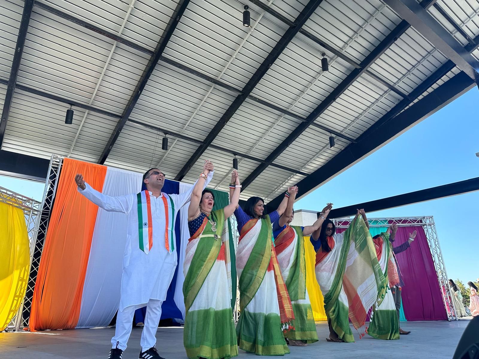 Group in traditional Indian attire on stage, arms raised. Behind them are colorful fabric panels under a metal roof.