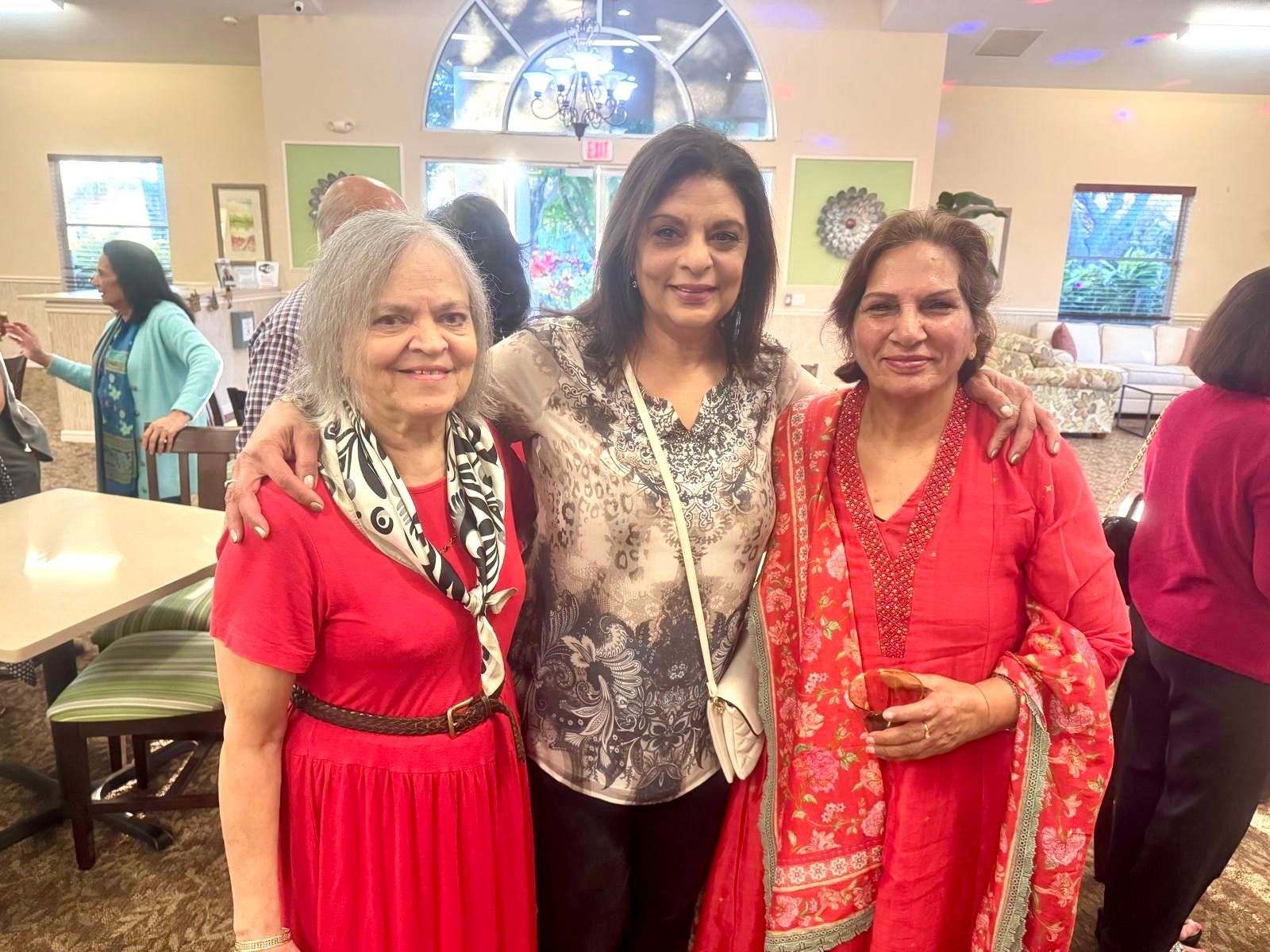 Three women smile, arm in arm, indoors. One in red, one in patterned top, one in red.
