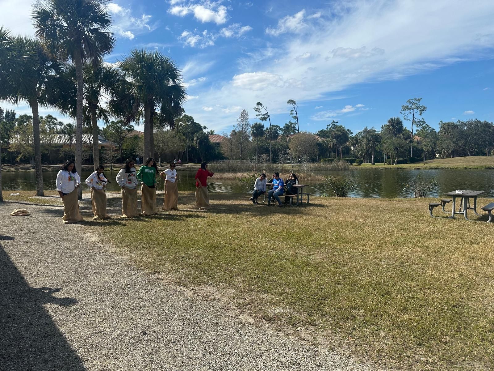 People playing sack race and others at a table near a lake on a sunny day.