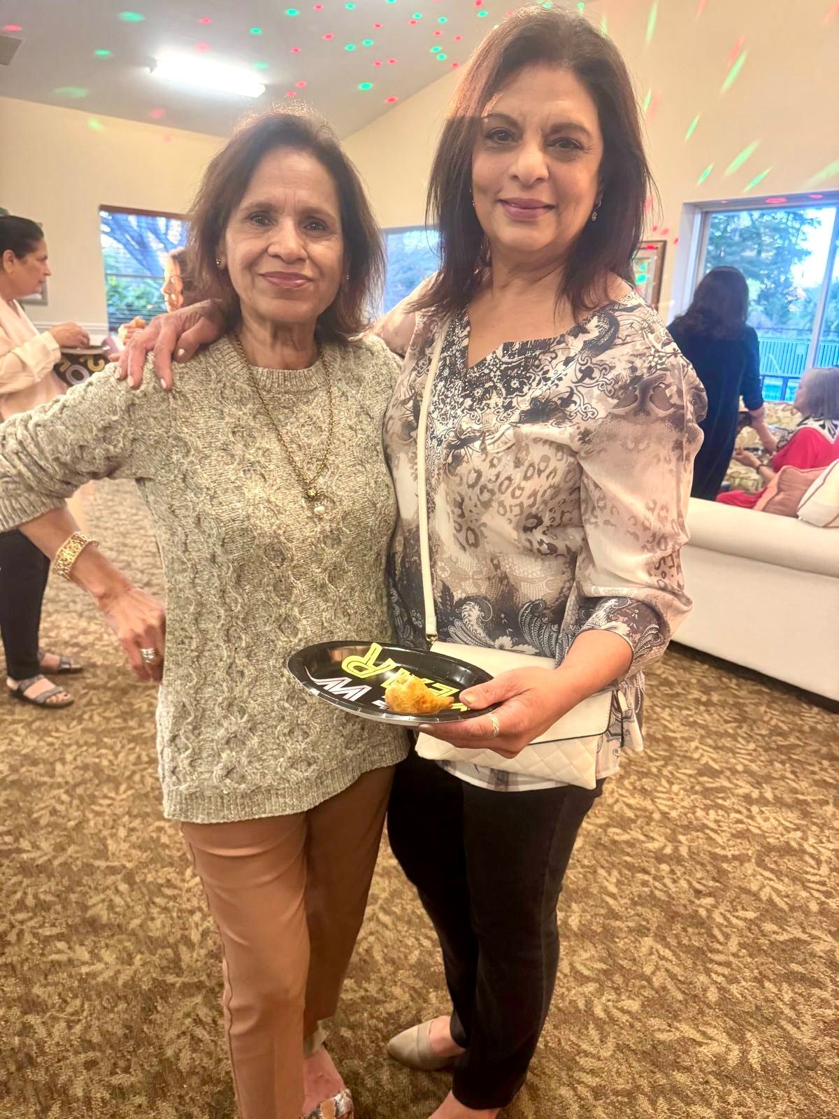 Two women pose together at a party. One holds food, the other has her arm around her. Indoors, smiling.