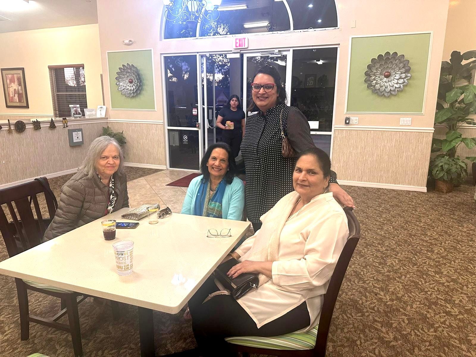 Four women pose around a table inside. One woman stands, smiling. Other women smile, one has a hand resting on the shoulder of the standing woman.