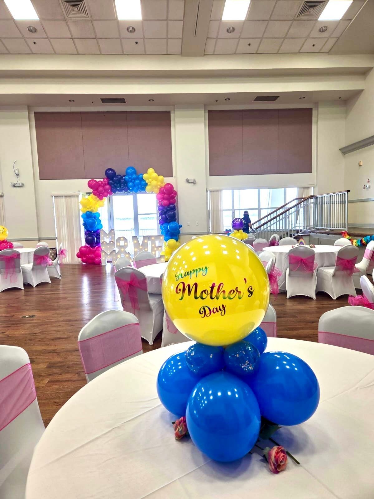 Mother's Day celebration with balloon centerpiece, blue and yellow balloons, tables, archway, indoor setting.