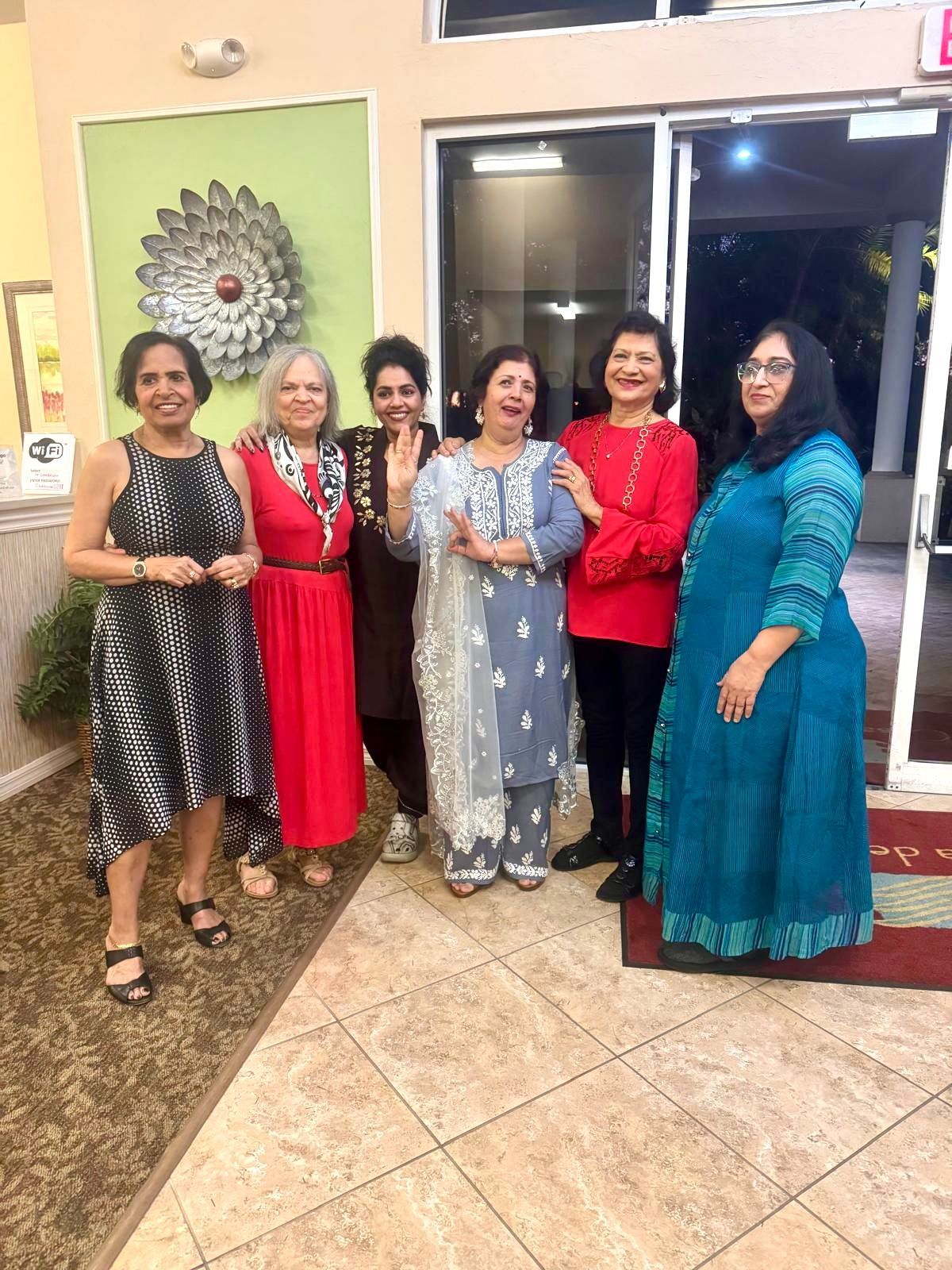 Six women pose indoors. One in a black dress, another in red, a third in brown, the next in a blue outfit, then red, and finally, a blue dress.
