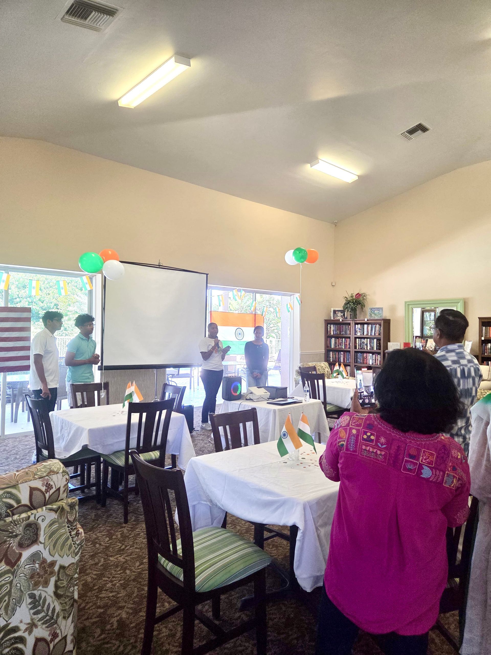 People celebrating India's Independence Day indoors. Tables are decorated with flags, balloons, and a screen for presentation.