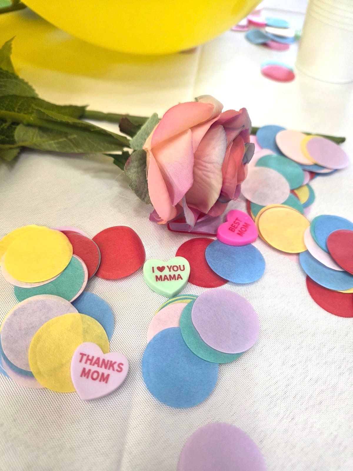 Rose, confetti, and conversation hearts on a white surface. Confetti colors: red, yellow, blue, purple, and green.