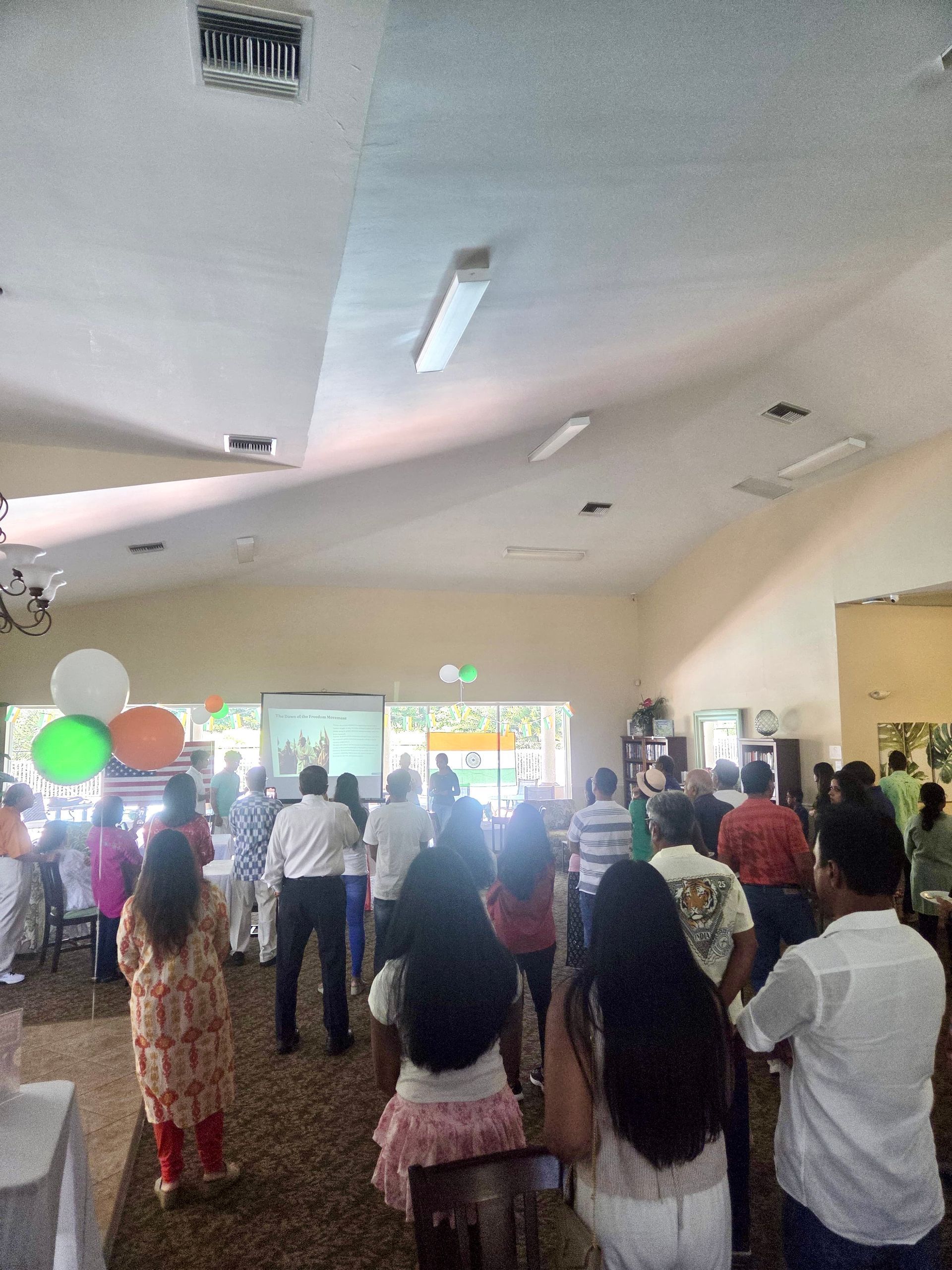 People gathered indoors, watching a presentation on a screen, with tricolor balloons nearby.