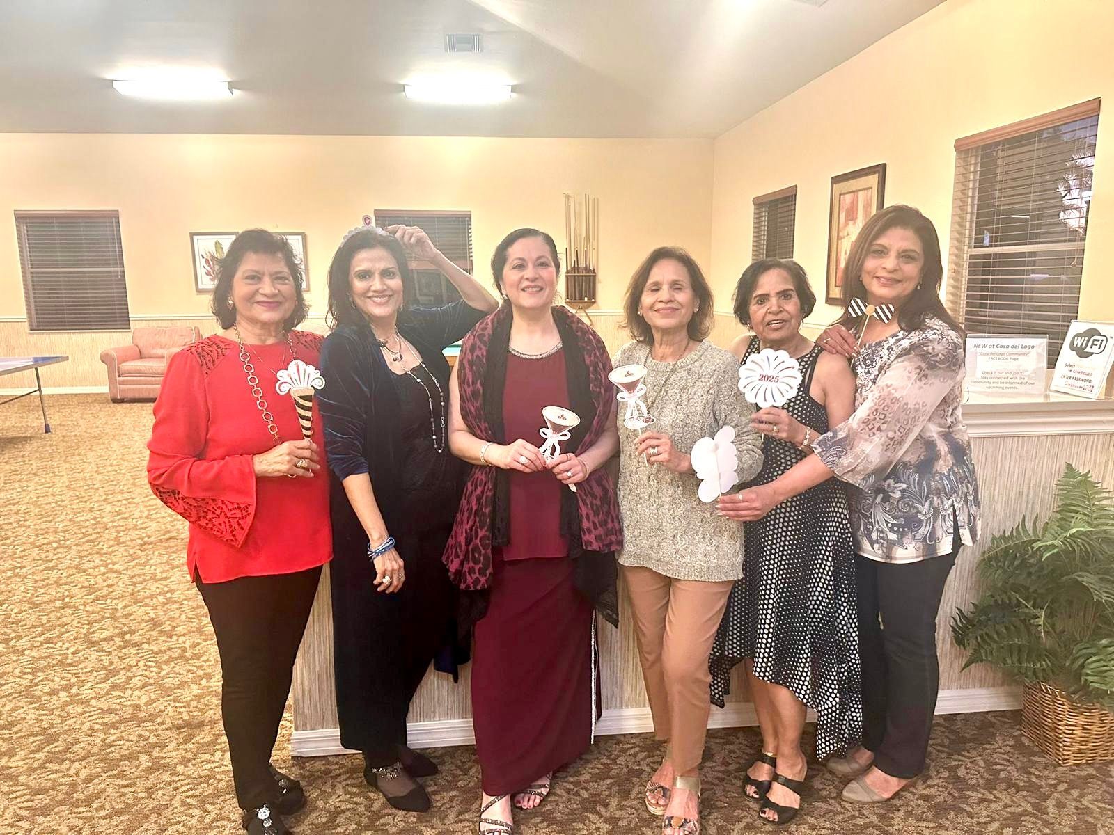 Six women pose holding flowers, indoors. They are smiling and wearing various elegant outfits.