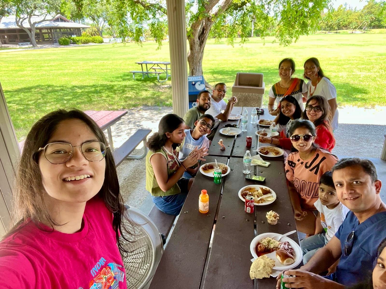 Group of people at picnic table, eating outdoors. Smiling faces, food on plates, park setting.