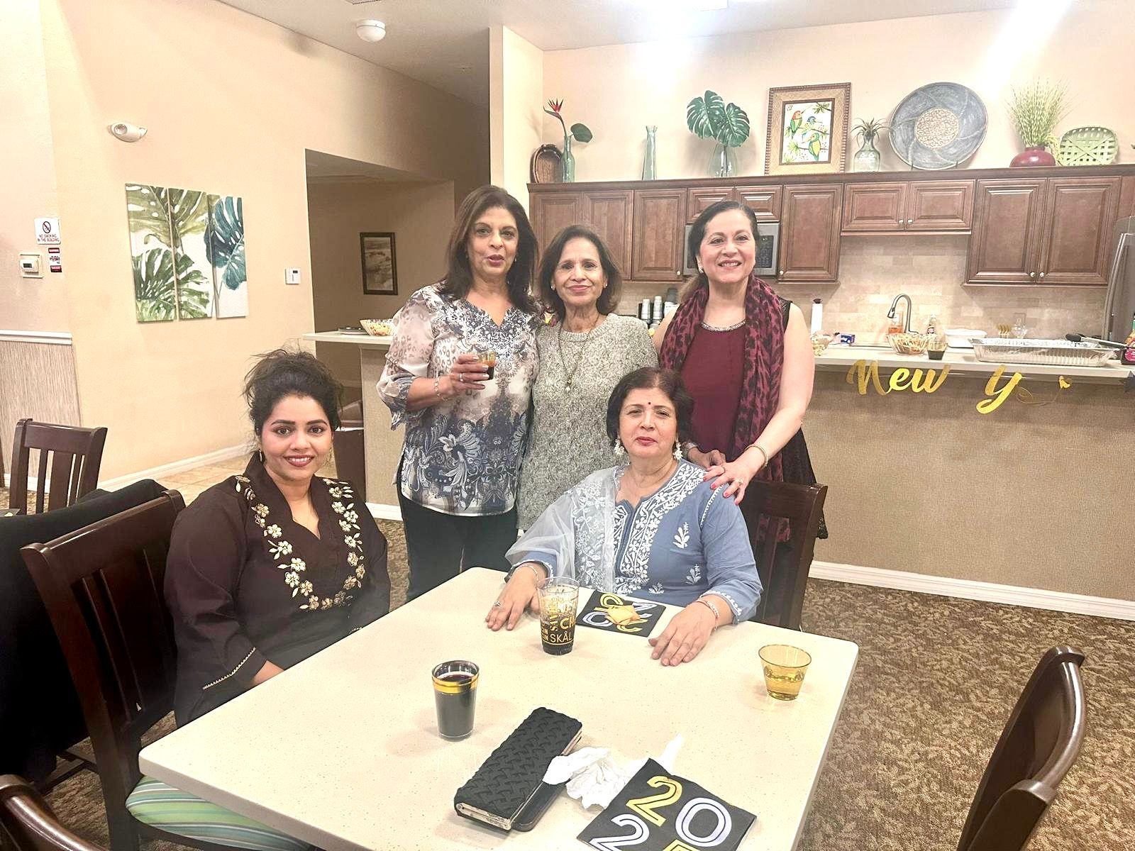 Five women at a table celebrating; beige walls, decor behind, dark hair, drinks.