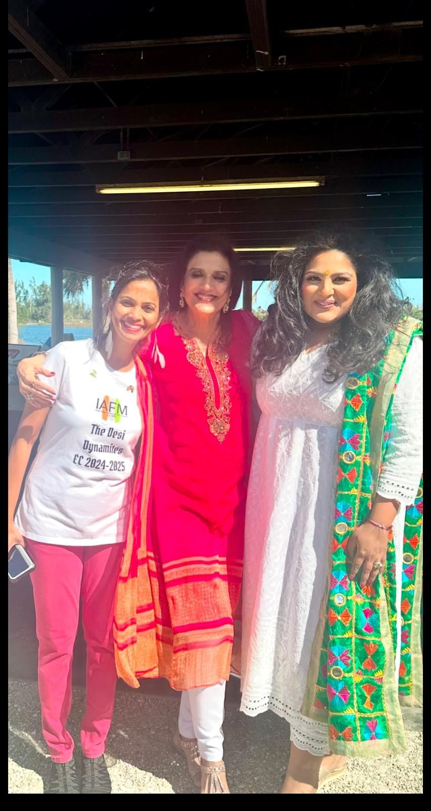 Three women smiling together outdoors. Two wear colorful ethnic clothing, one in a white t-shirt and pink pants.
