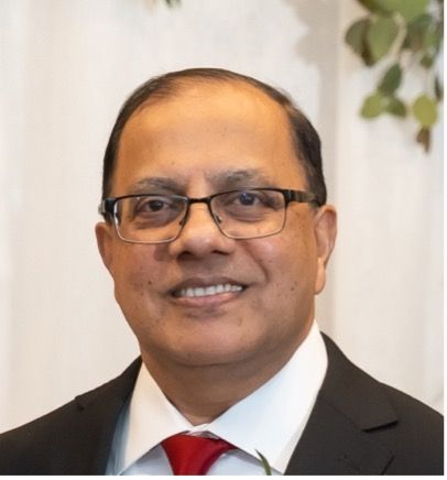 Man in suit with glasses smiling. Red tie, blurred floral background.