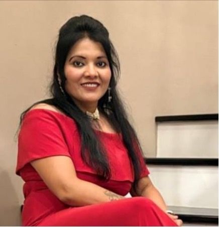 Woman with dark hair in a red off-the-shoulder top smiling, seated near stairs.