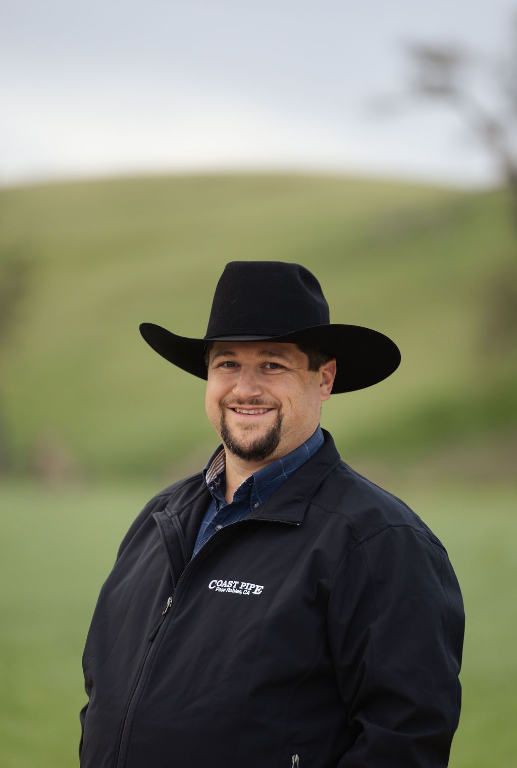 Smiling person in a black cowboy hat and jacket standing outdoors on a grassy hill