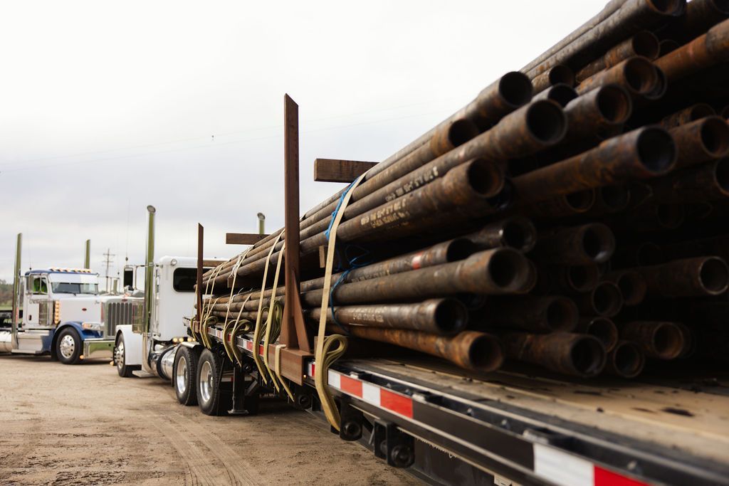 A white semi-truck hauls a flatbed trailer stacked with a large load of weathered, rusted metal pipes.