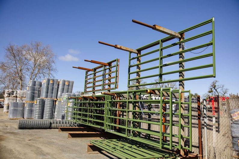 Stacked green metal livestock panels and rolls of wire fencing in an outdoor yard under a clear blue sky.