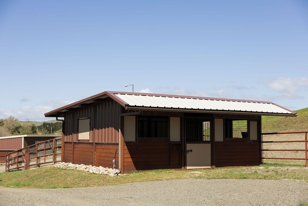 Small brown wooden building with a metal white roof in a fenced gravel yard under a clear sky