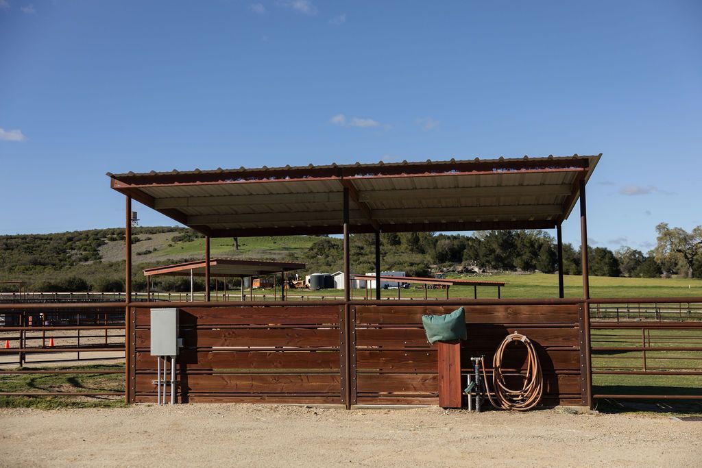Rusty livestock pen with a metal-roofed shelter, fencing, and a coiled hose in a dusty ranch yard.