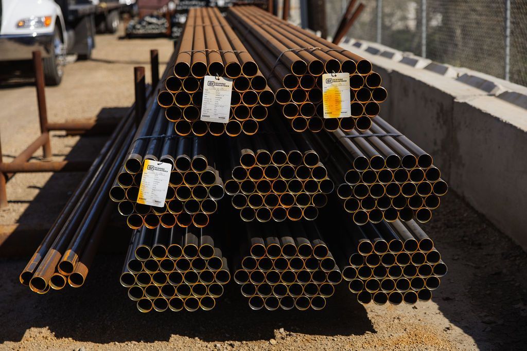 Stacks of black and rusty steel pipes with tags, piled outdoors on a gravel lot near a road barrier.
