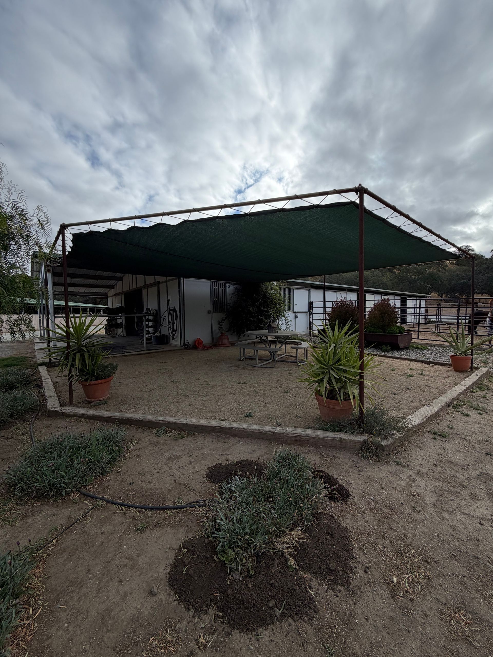 Outdoor patio with a green canopy, picnic tables, potted plants, and a cloudy sky.