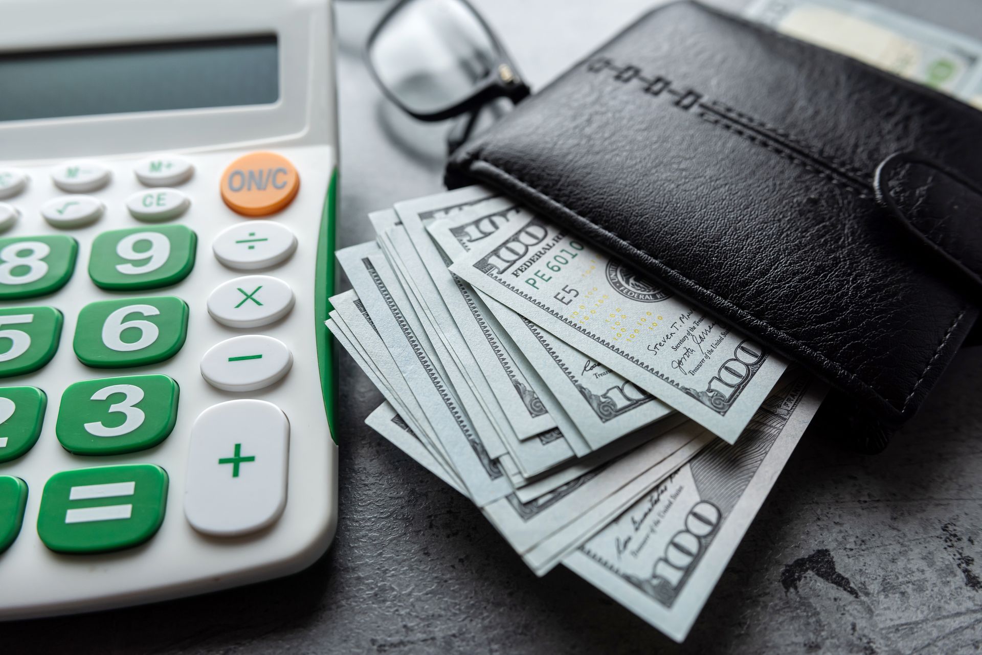 Calculator and black wallet overflowing with cash on a gray surface.