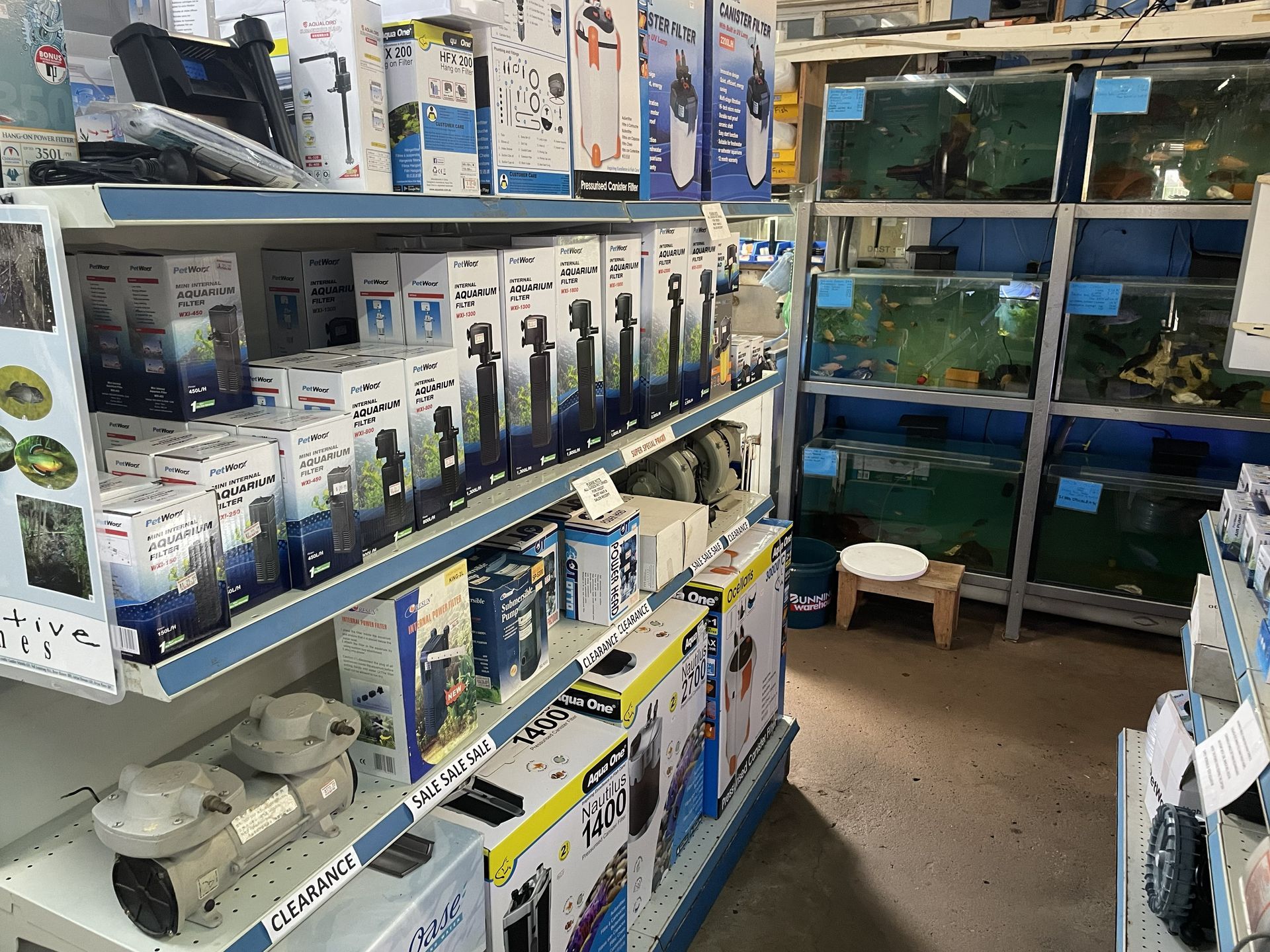 Shelves of aquarium supplies in a pet store, tanks with fish in background.