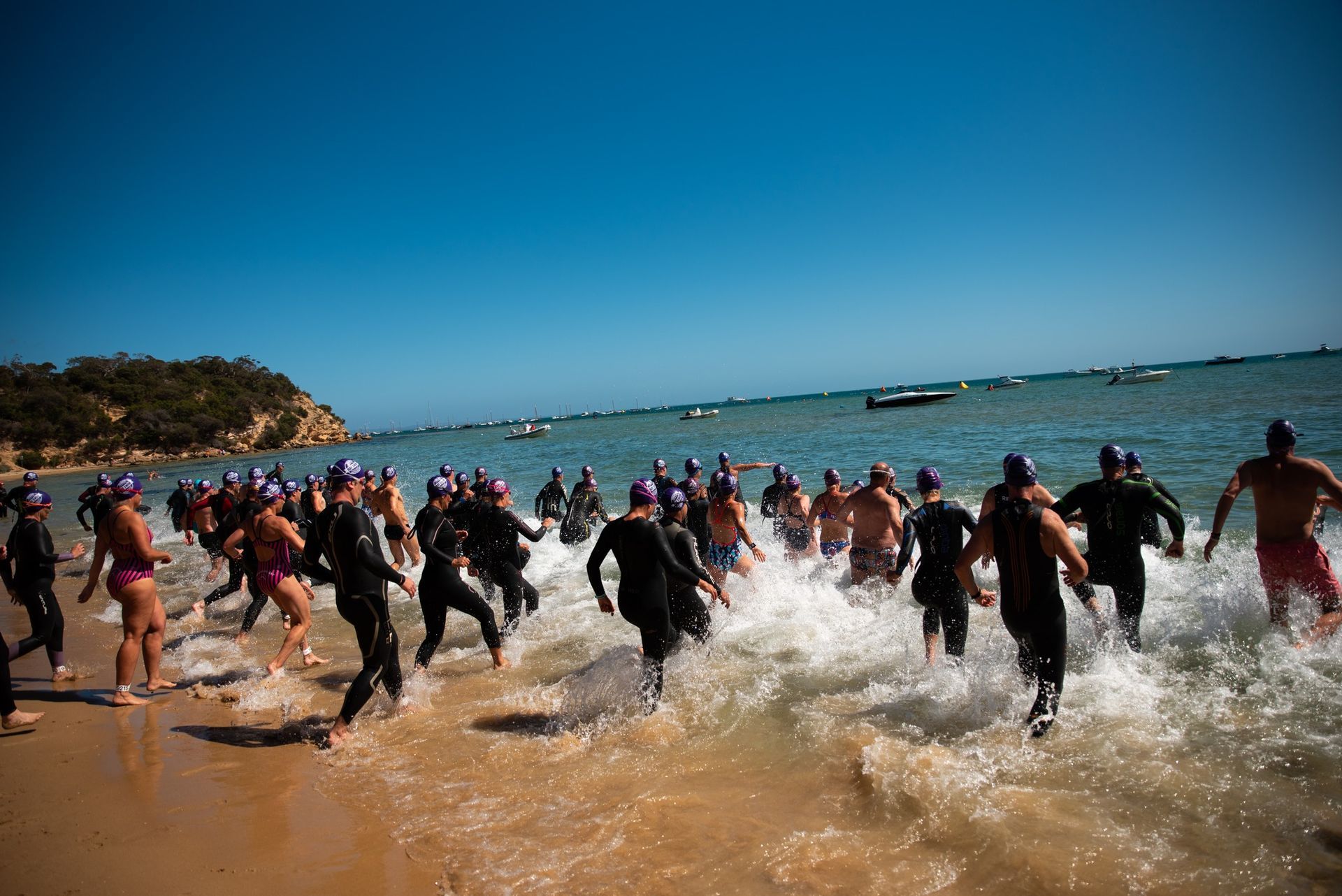 Triathletes running into ocean, waves crashing on shore. Blue sky, sunny day.