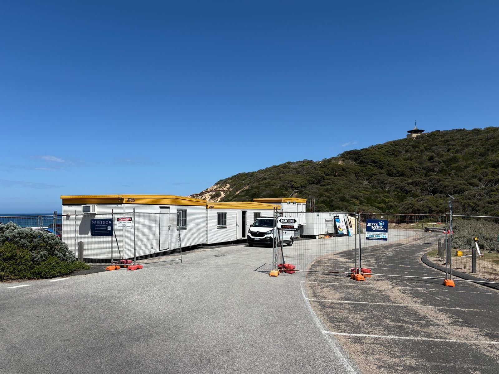 Construction trailers on a gravel lot near a hill and water. A vehicle is parked in front. Blue sky.