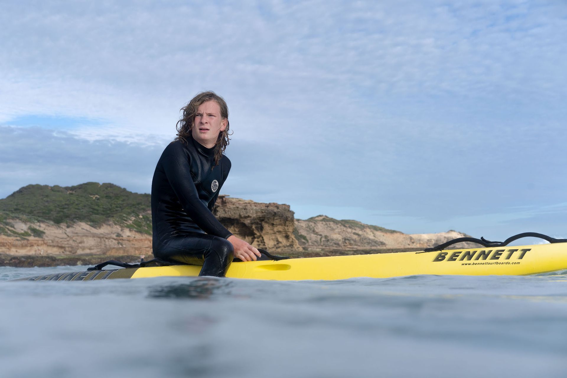 Person in wetsuit on a yellow rescue board in the ocean, rocky coast in background.