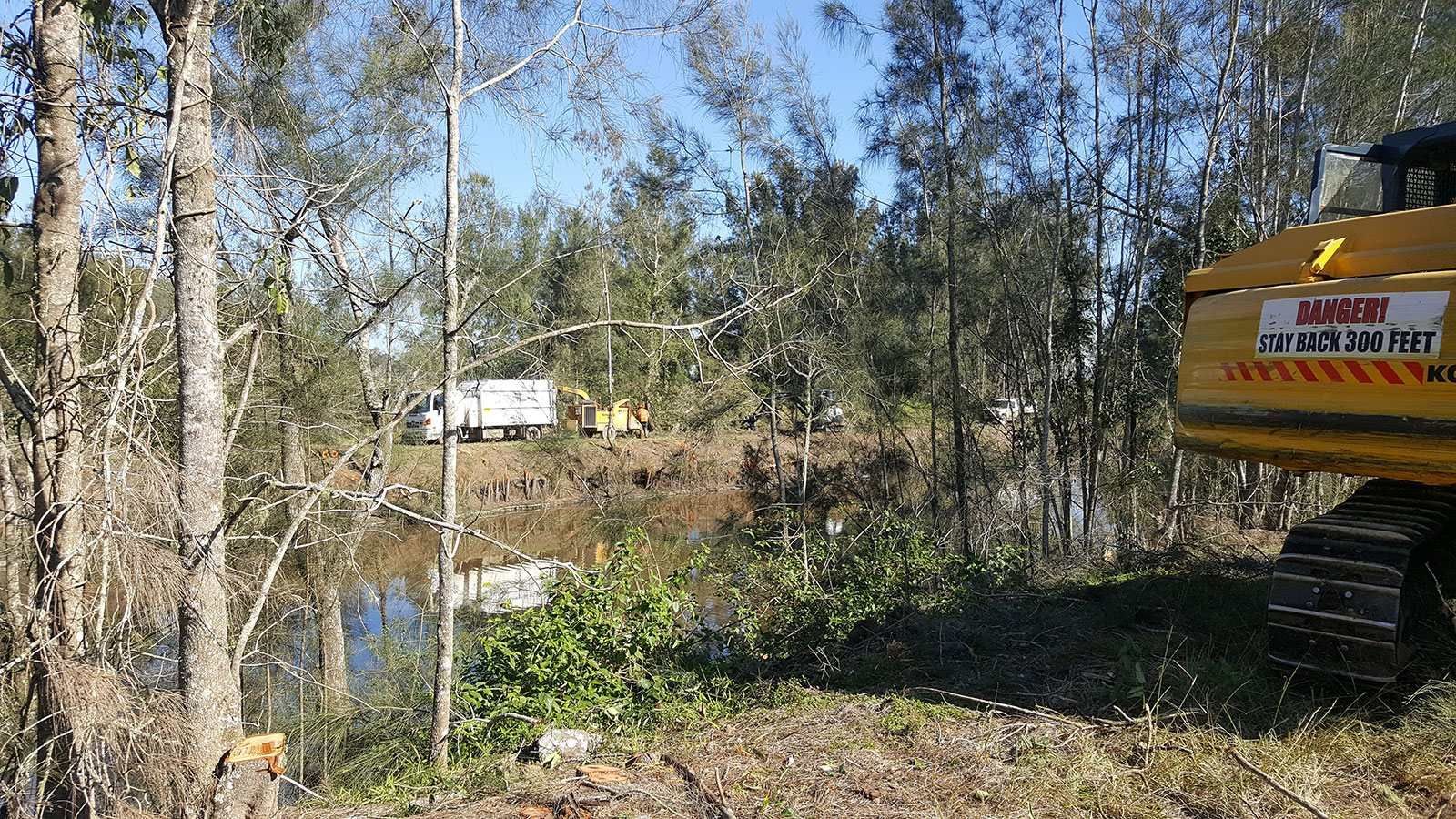 A yellow excavator is parked in the middle of a forest next to a body of water.