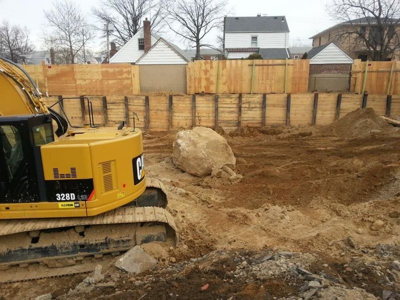 Yellow excavator at a construction site with a large rock, surrounded by wooden fencing and dirt.
