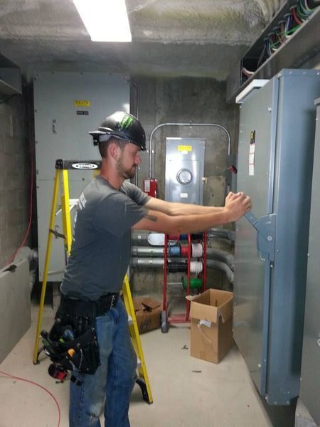 Electrician in a gray shirt and jeans opening an electrical panel in a utility room.