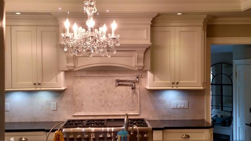 Cream-colored kitchen with range, range hood, cabinets, chandelier, dark countertops, and neutral backsplash.