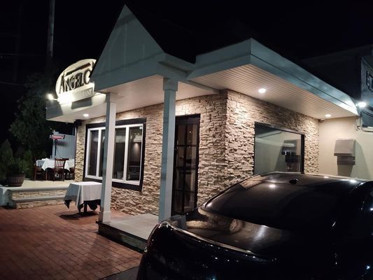 Restaurant exterior at night with tan brick facade, black car in front, and outdoor seating.