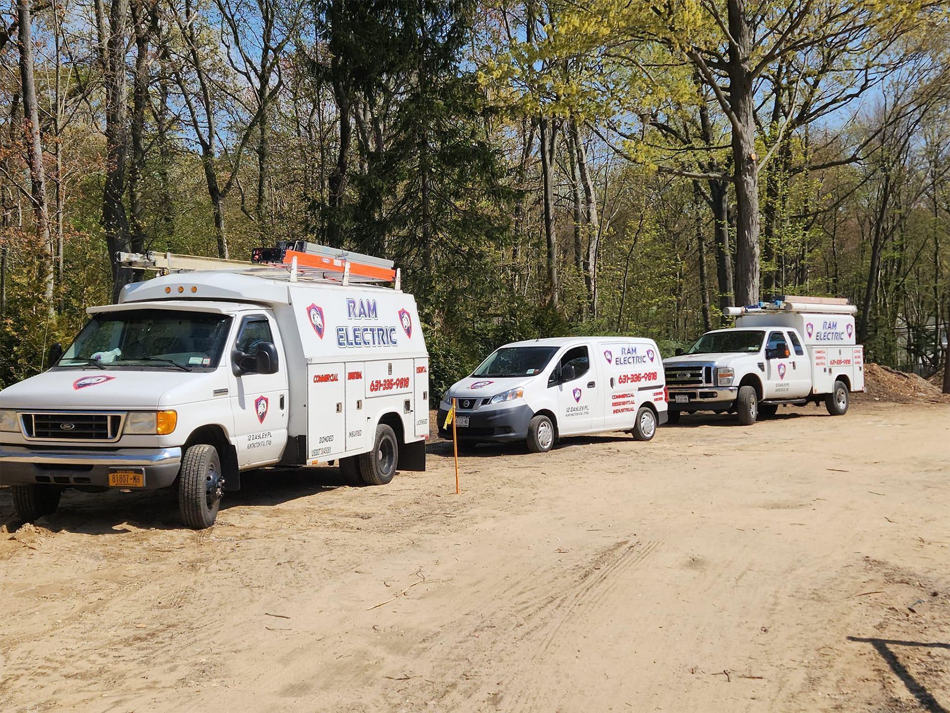 Three white utility trucks parked on a dirt road in a wooded area;