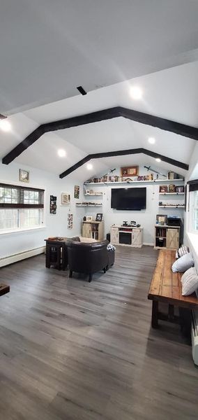 Living room with a vaulted ceiling, black beams, and gray flooring. There's a couch and TV, with windows on the left.