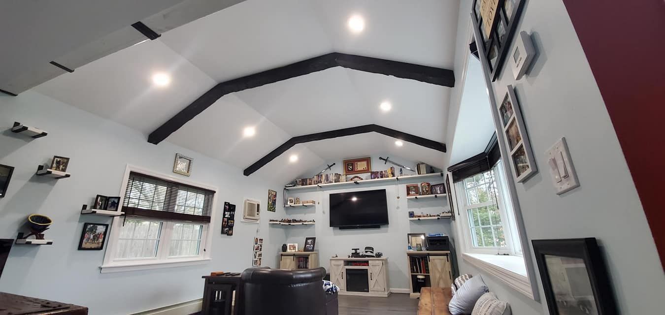 A cozy living room with white walls, dark beams, and a TV, surrounded by windows, shelves, and framed photos.