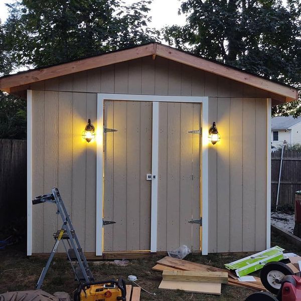 Tan shed with double doors, outdoor lights, and tools on the ground.