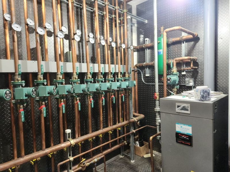 Copper pipes and valves in a mechanical room, with a gray water heater and various gauges.