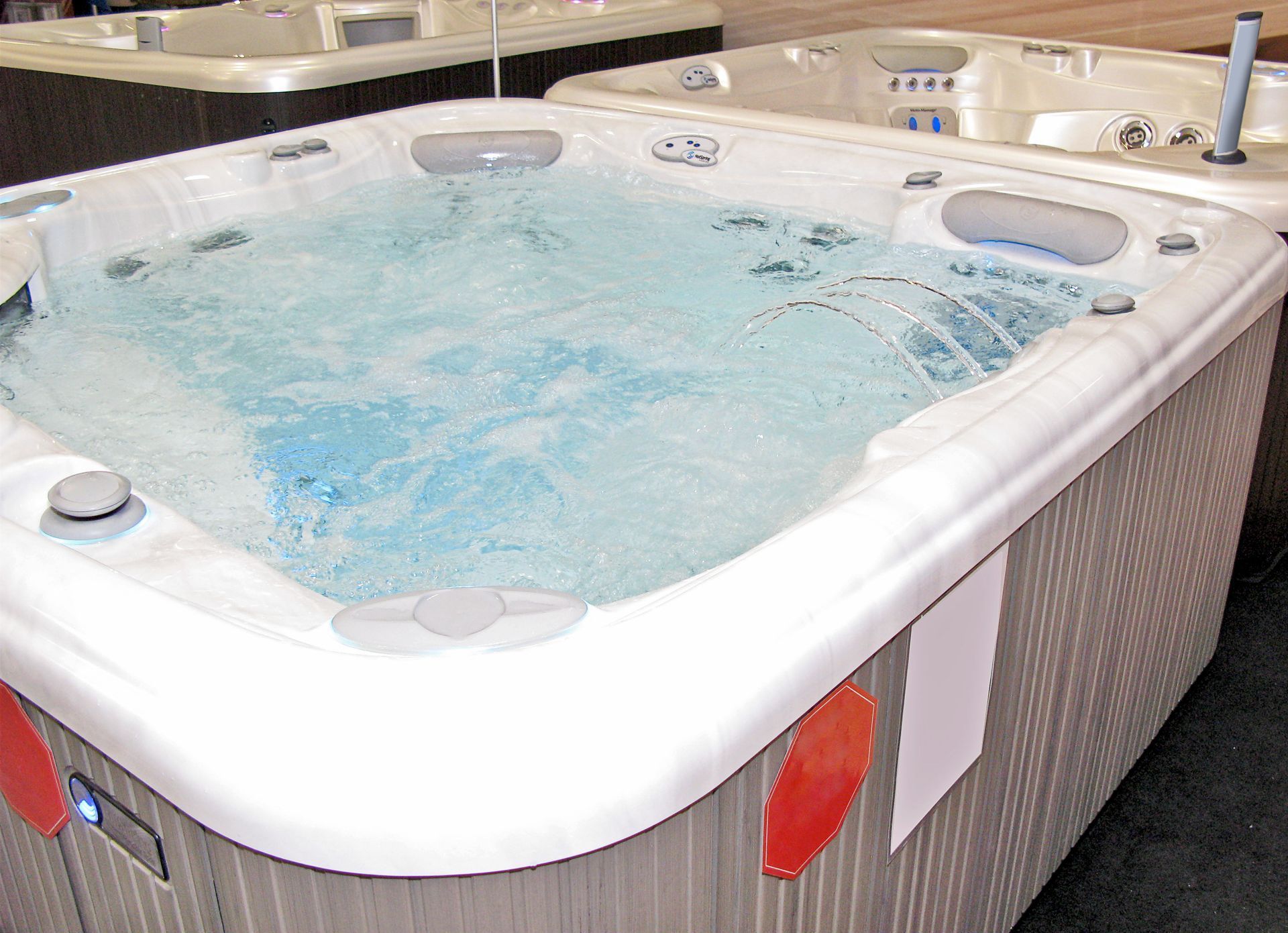 A square, white hot tub filled with blue water, with jets spraying. Grey-brown siding visible.