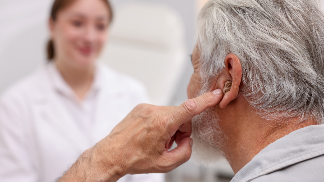 An older person adjusting a hearing aid in their ear while facing a medical professional in a bright clinical setting.