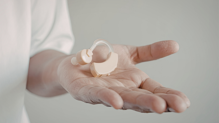 A beige behind-the-ear hearing aid resting on an open palm against a plain, light background.