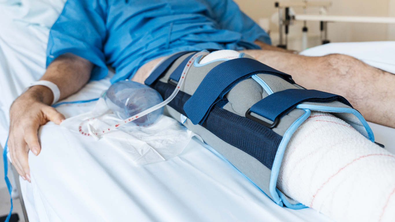 A patient in a hospital bed with a bandaged leg secured in a knee brace and a cryotherapy cooling wrap.