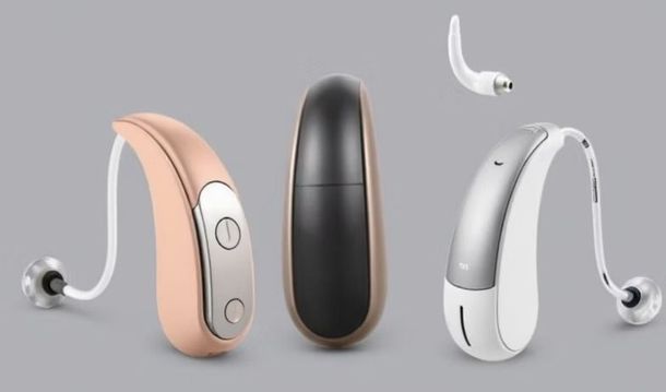 Three hearing aids in rose gold, black, and white-silver, featuring thin tubes and ear domes against a gray background.