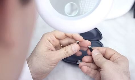 A person uses a magnifying lamp to examine small jewelry components held in their hands over a small storage case.