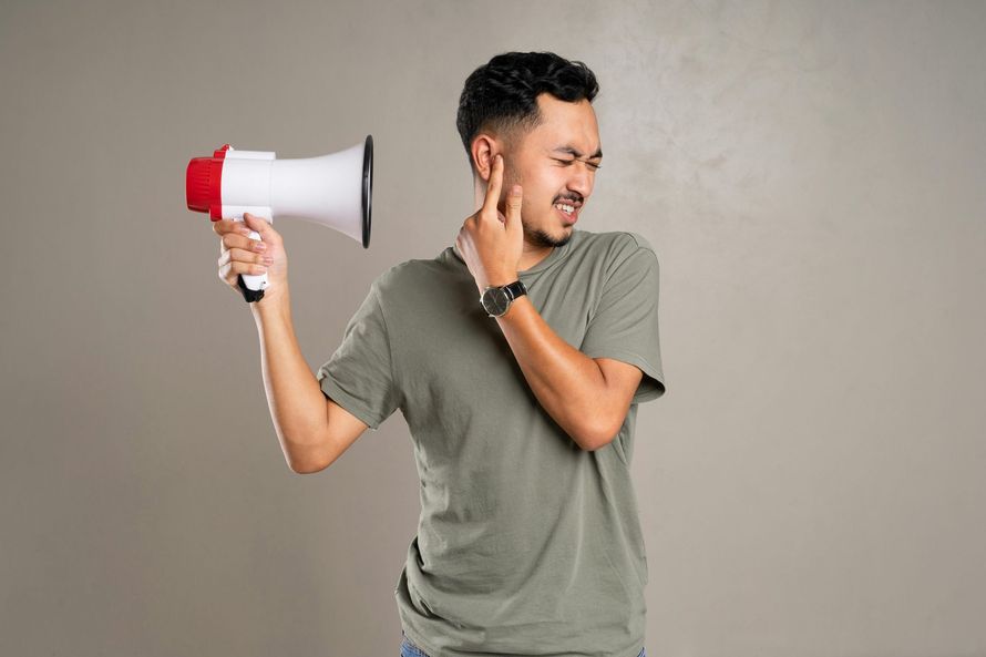 A person in a gray t-shirt winces while holding a megaphone near their ear, covering it with their finger.