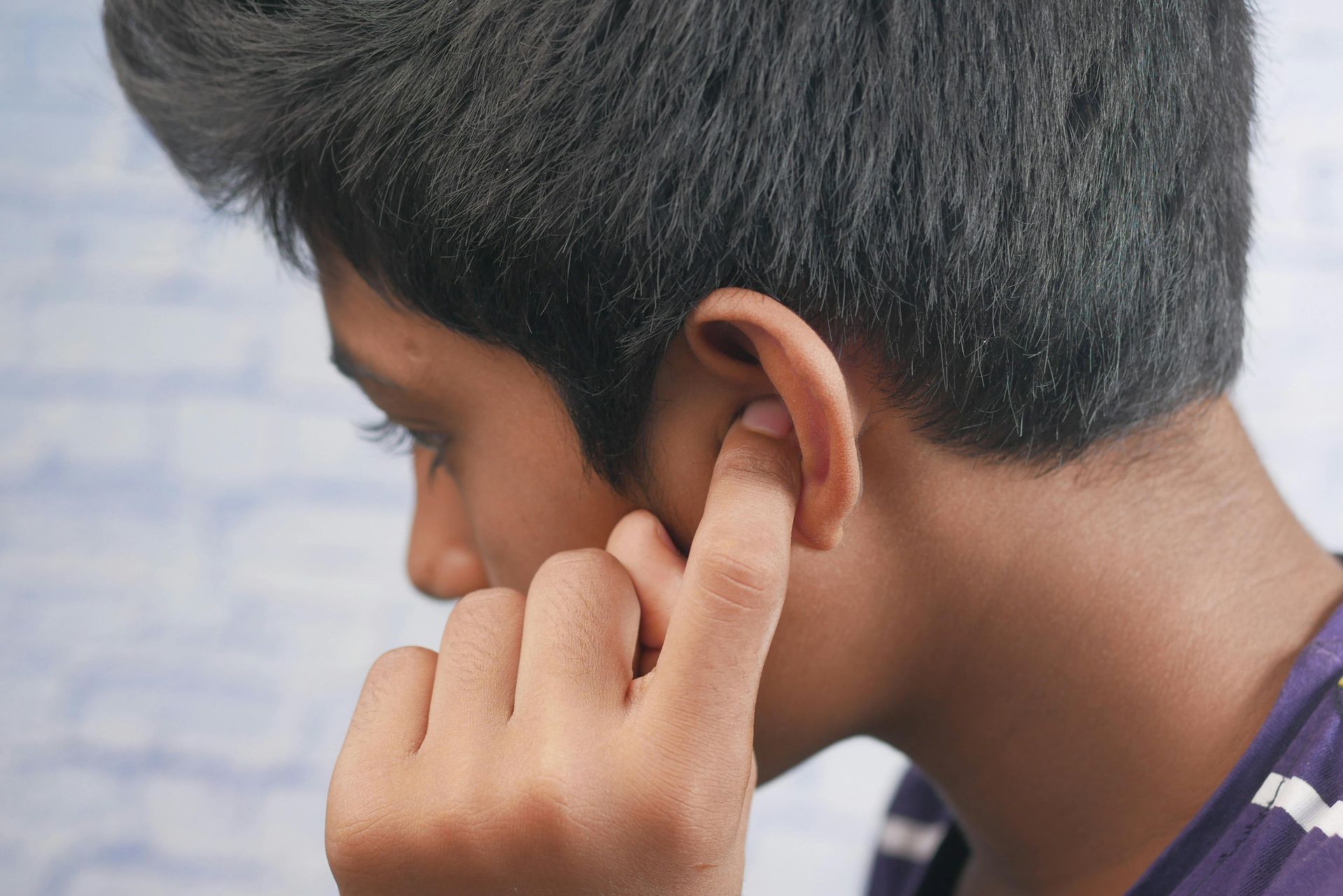 A person using a finger to scratch or clean the inside of their ear against a light blue, brick-patterned background.