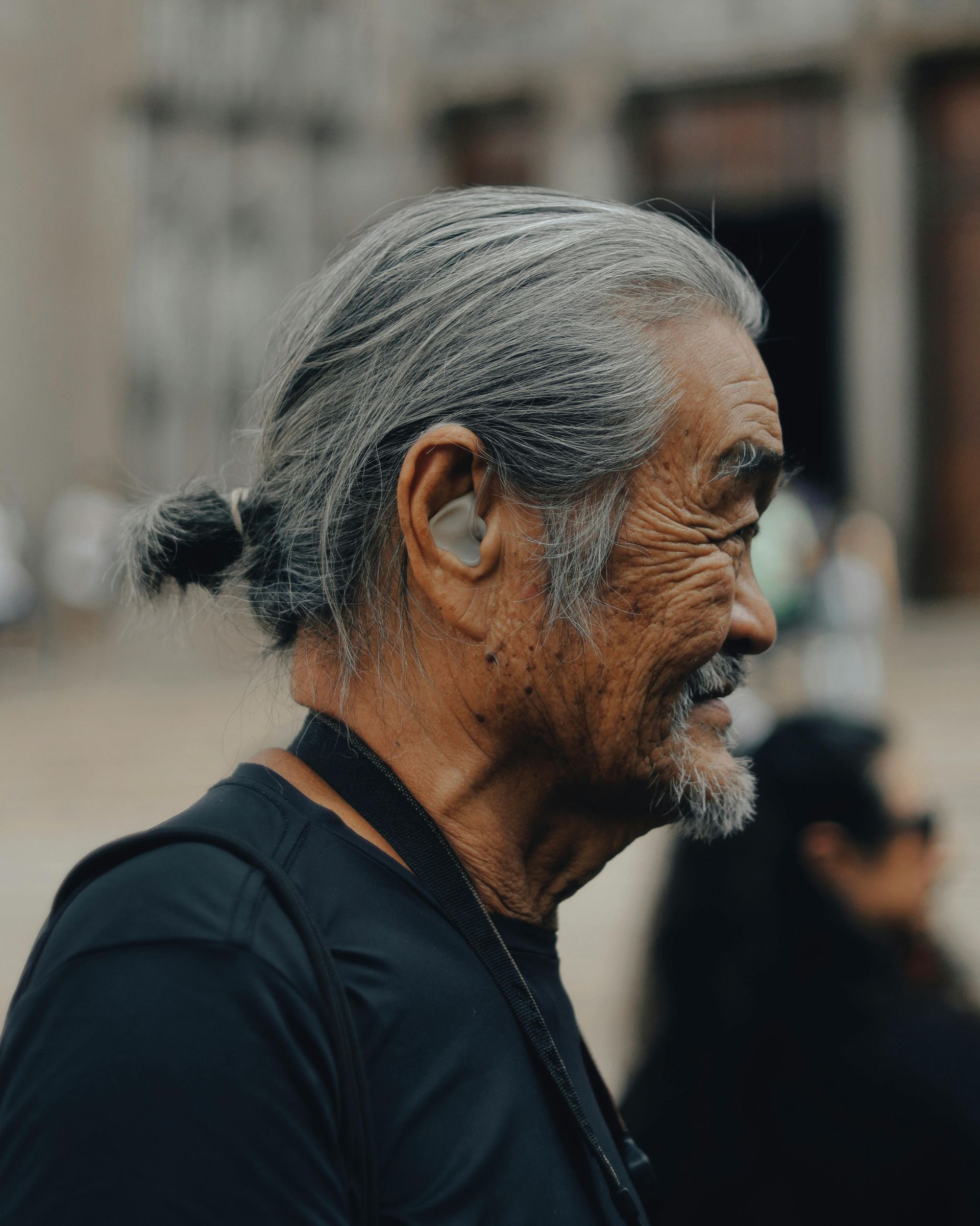 A side profile of an individual with graying hair tied in a small bun, wearing a black shirt in an outdoor setting.