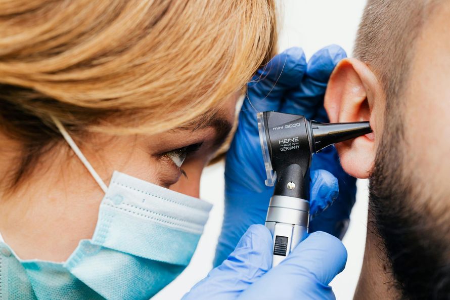A medical professional wearing a mask and blue gloves examines a patient's ear using an otoscope.